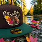 A dark brown and green New Era 59FIFTY fitted baseball cap with an embroidered moose head and autumn leaves design on the front, resting on a log outdoors with fallen leaves and a forest lake in the background.