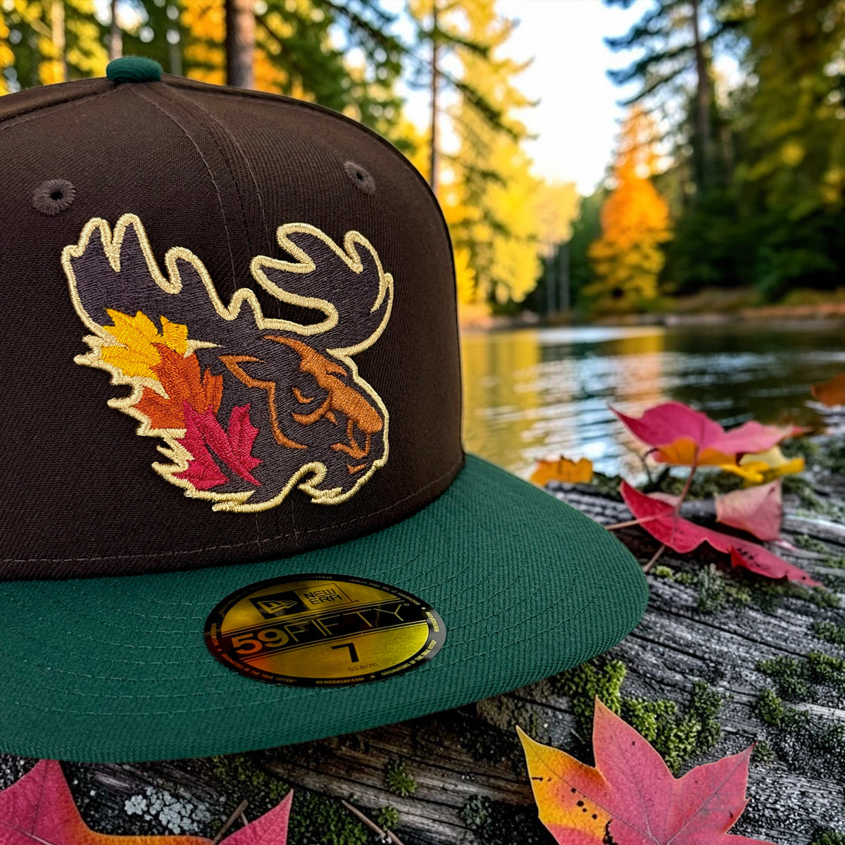 A dark brown and green New Era 59FIFTY fitted baseball cap with an embroidered moose head and autumn leaves design on the front, resting on a log outdoors with fallen leaves and a forest lake in the background.