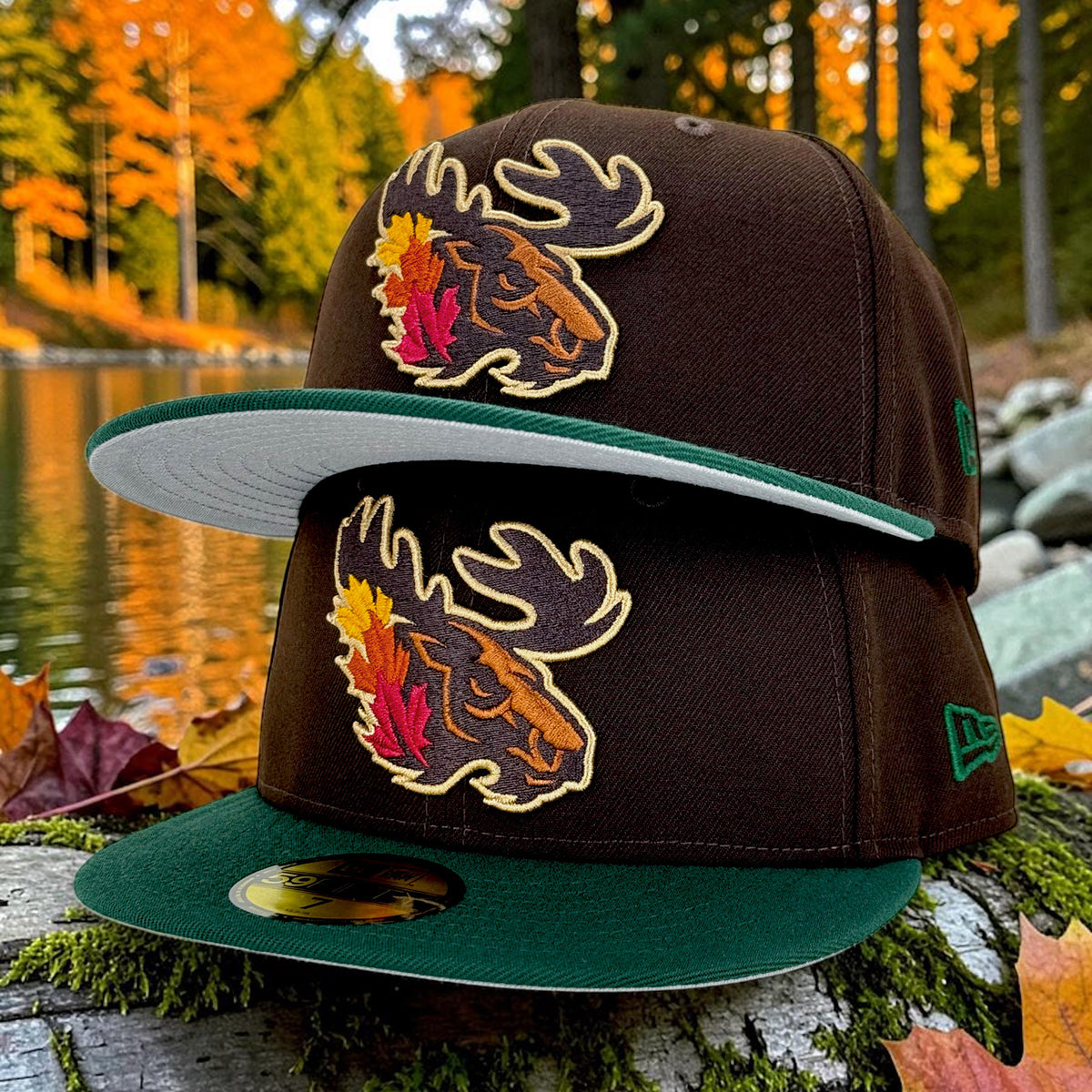 Two dark brown fitted caps with green visors are stacked on a mossy log outdoors near a lake, with autumn trees in the background. Each cap has a large embroidered moose head logo with colorful fall leaves and gold outlining, and a small green New Era flag logo is visible on the side.